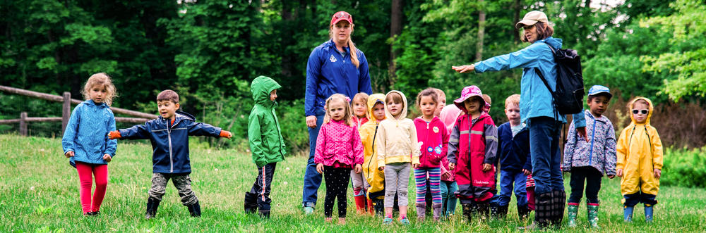 Students and teachers at Drumlin Farm Community Preschool lined up outdoors in a grassy field, preparing to play a game