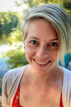 A portrait photo of Hilary Johansen-Silve with green trees and still water in the background. She has short, blond hair and is wearing a red blouse and white sweater.