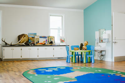 A photo of the inside of the Stony Brook Nature Preschool classroom, with small chairs and a table, a rug with a pond design, and various nature items and books on a shelf in the back