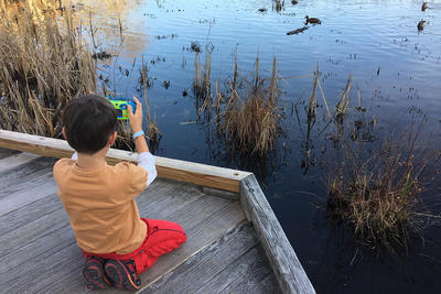 A young child kneeling on a wooden boardwalk holding a child's camera, pointed toward something in the water