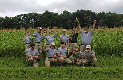 All 11 AmeriCorps members posing with vegetables in front of a field of corn