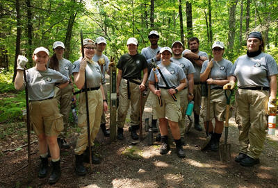 All 11 AmeriCorps members standing together on a trail in the woods holding varied trail building tools.