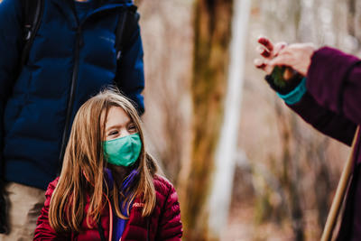A young girl wearing a face mask and puffy coat looks at a specimen held by an adult