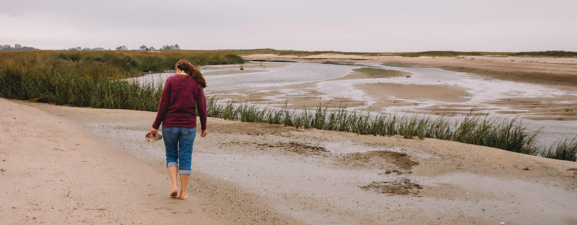 A person in long sleeves holding their sandals while walking barefoot on a sandy beach trail at Wellfleet Bay Wildlife Sanctuary