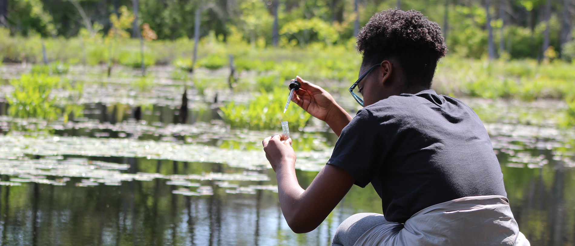 Student using a dropper to put water into a test tube, kneeling in front of a pond