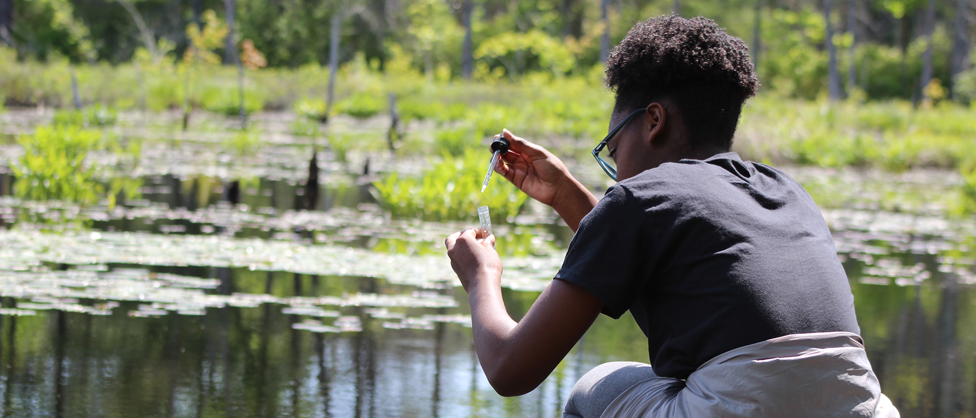 Student using a dropper to put water into a test tube, kneeling in front of a pond