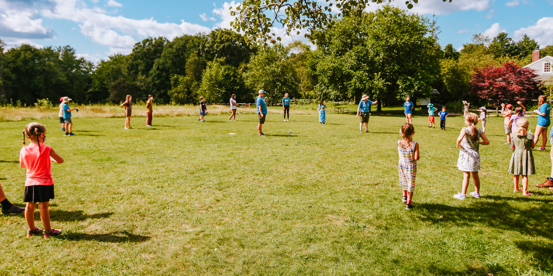 Campers at Stony Brook Nature Camp gather in a large circle on the activity field, listening to the camp directors in the middle explain the next game