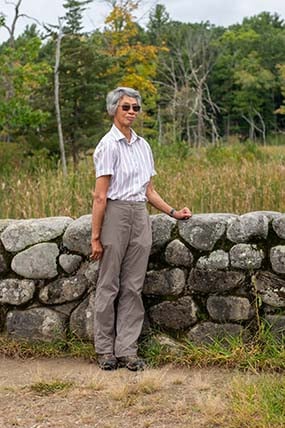 Woman with short gray hair, a button up shirt, and gray pants standing against a rock wall.