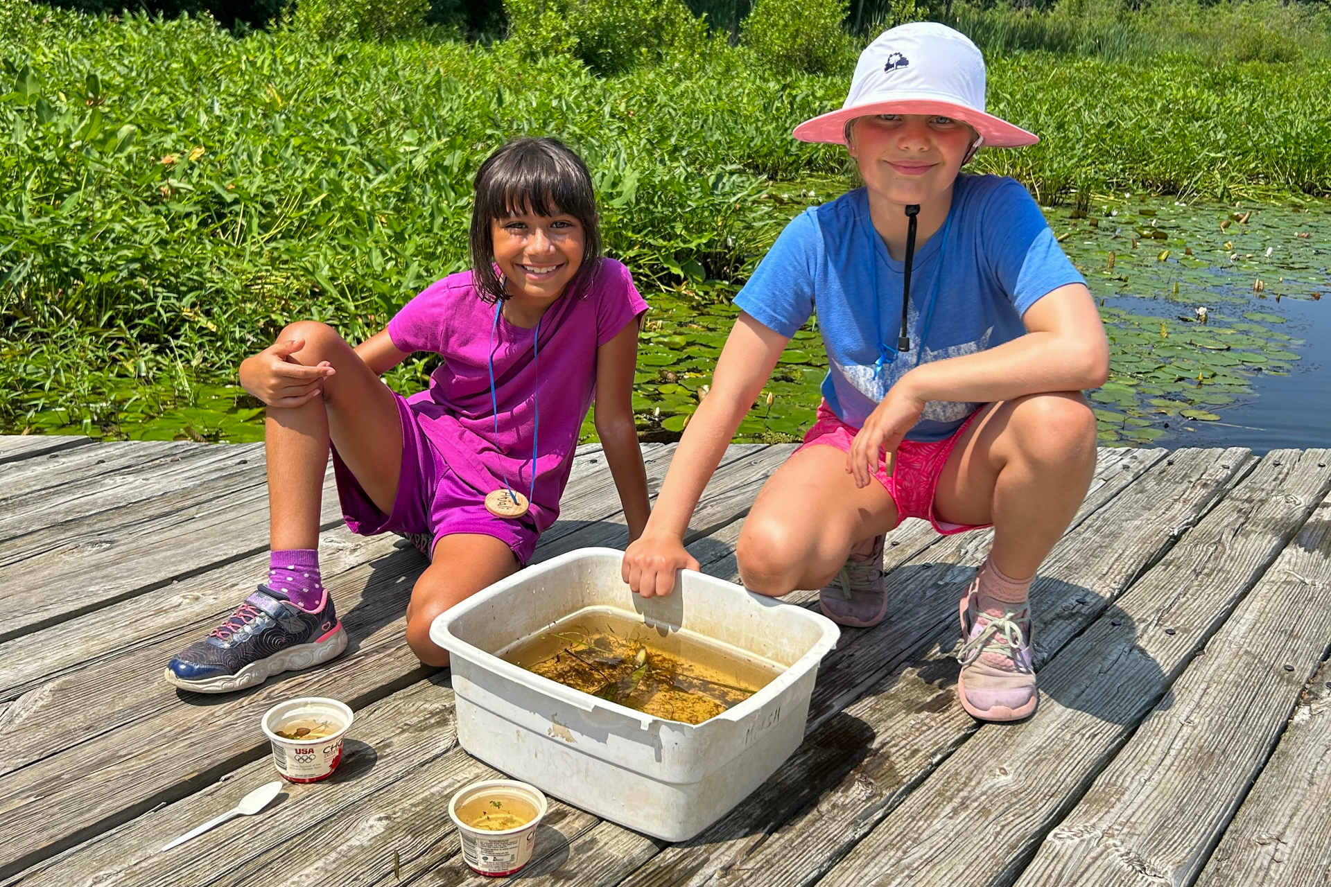 Two happy campers at Broadmoor Nature Camp kneeling behind a white bin filled with water, mud, and aquatic creatures