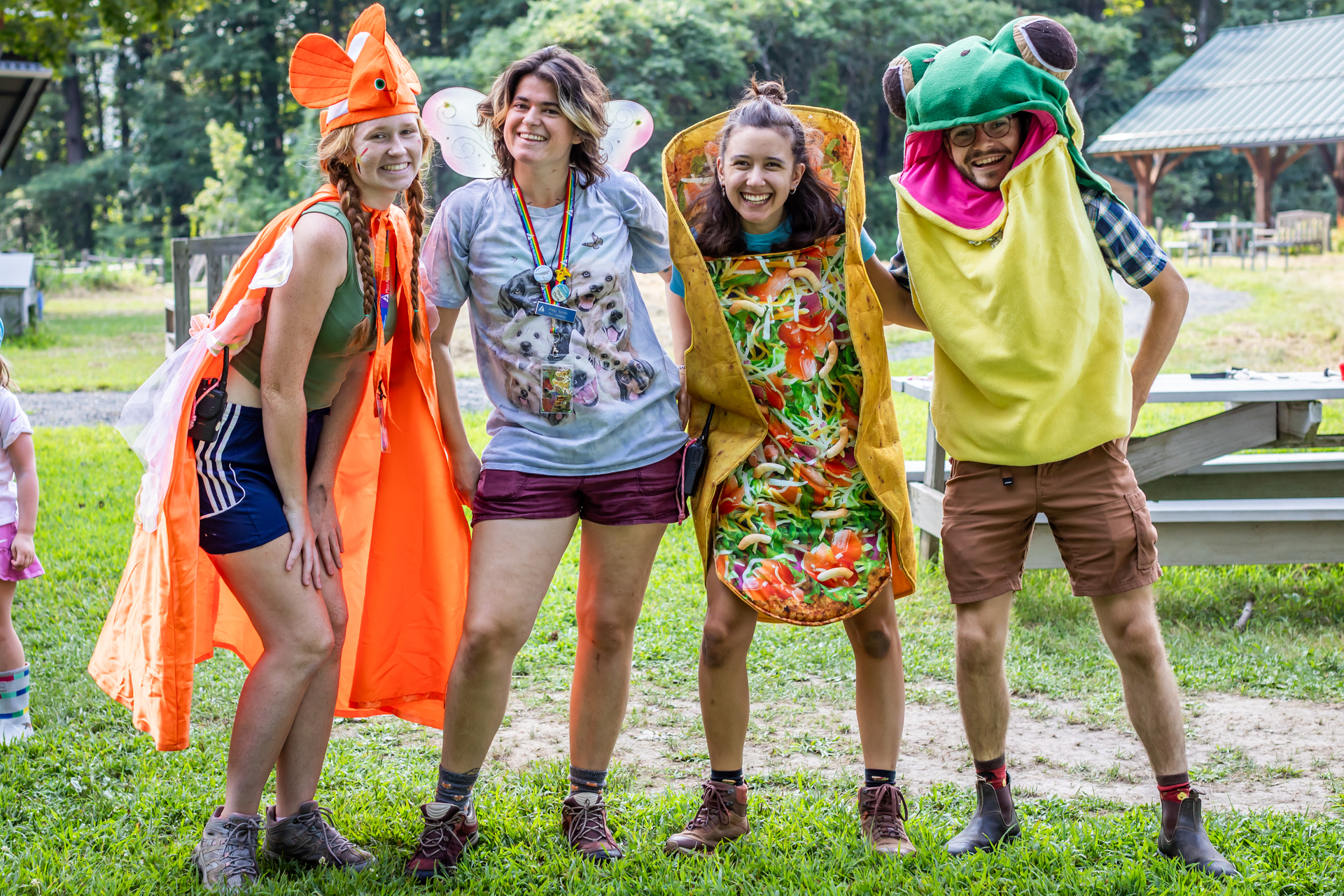Four counselors stand arm-in-arm wearing silly costumes- a fish, a fairy, a taco and a frog.