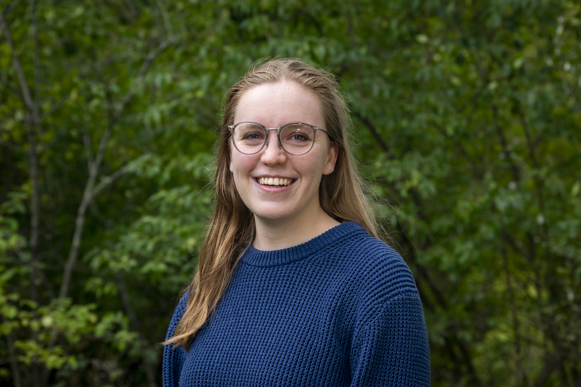 Anna is wearing a blue sweater and glasses, smiling with trees in the background.