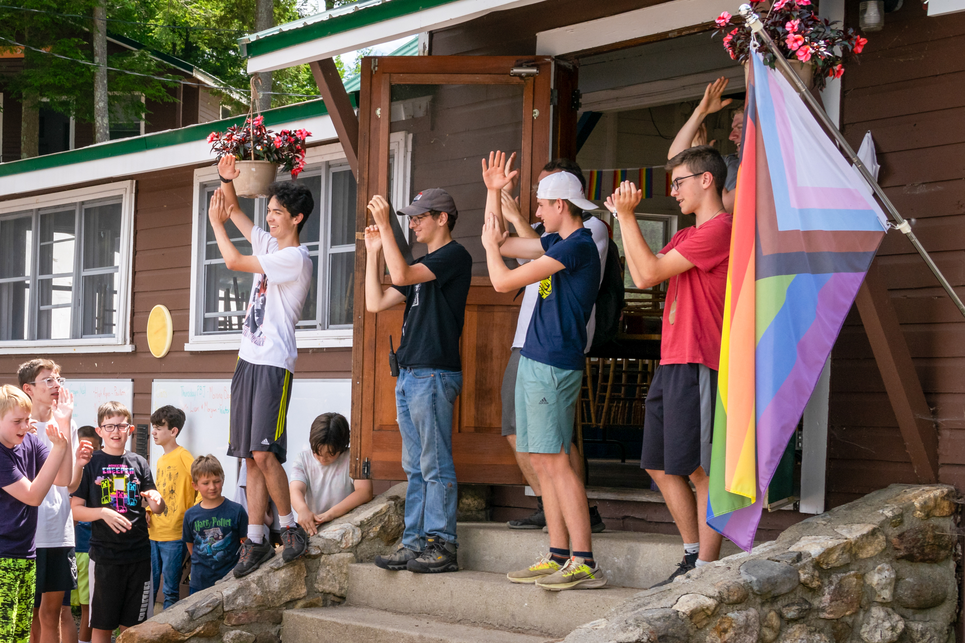 A group of Wildwood's Environmental Leadership Teens lead a camp song outside the dining hall; a Progress Pride flag flies in the foreground.