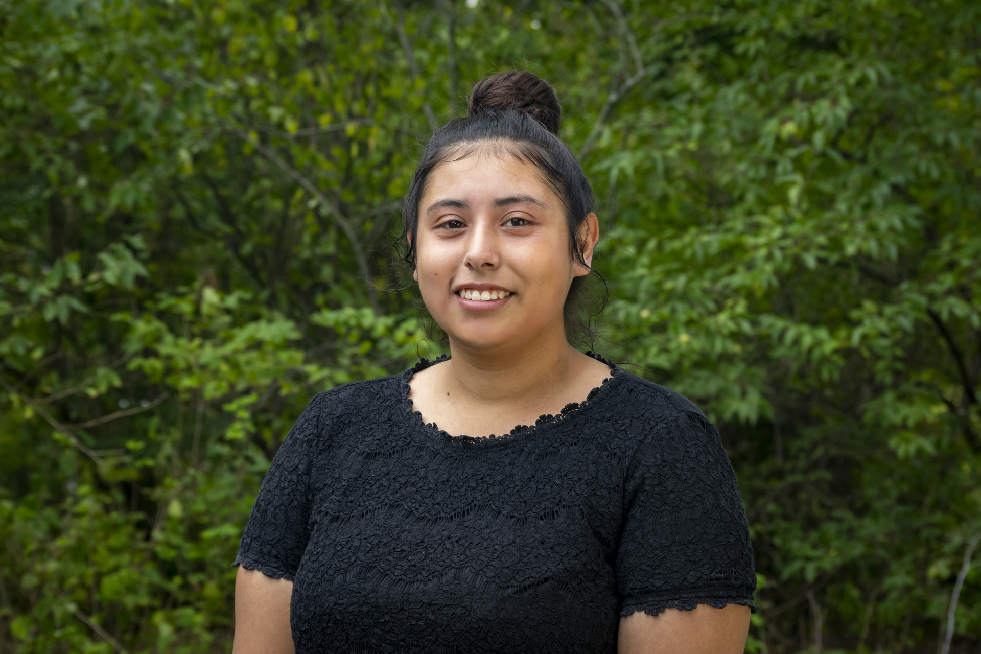 Isabela wearing a black shirt, smiling with green leaves in the background.