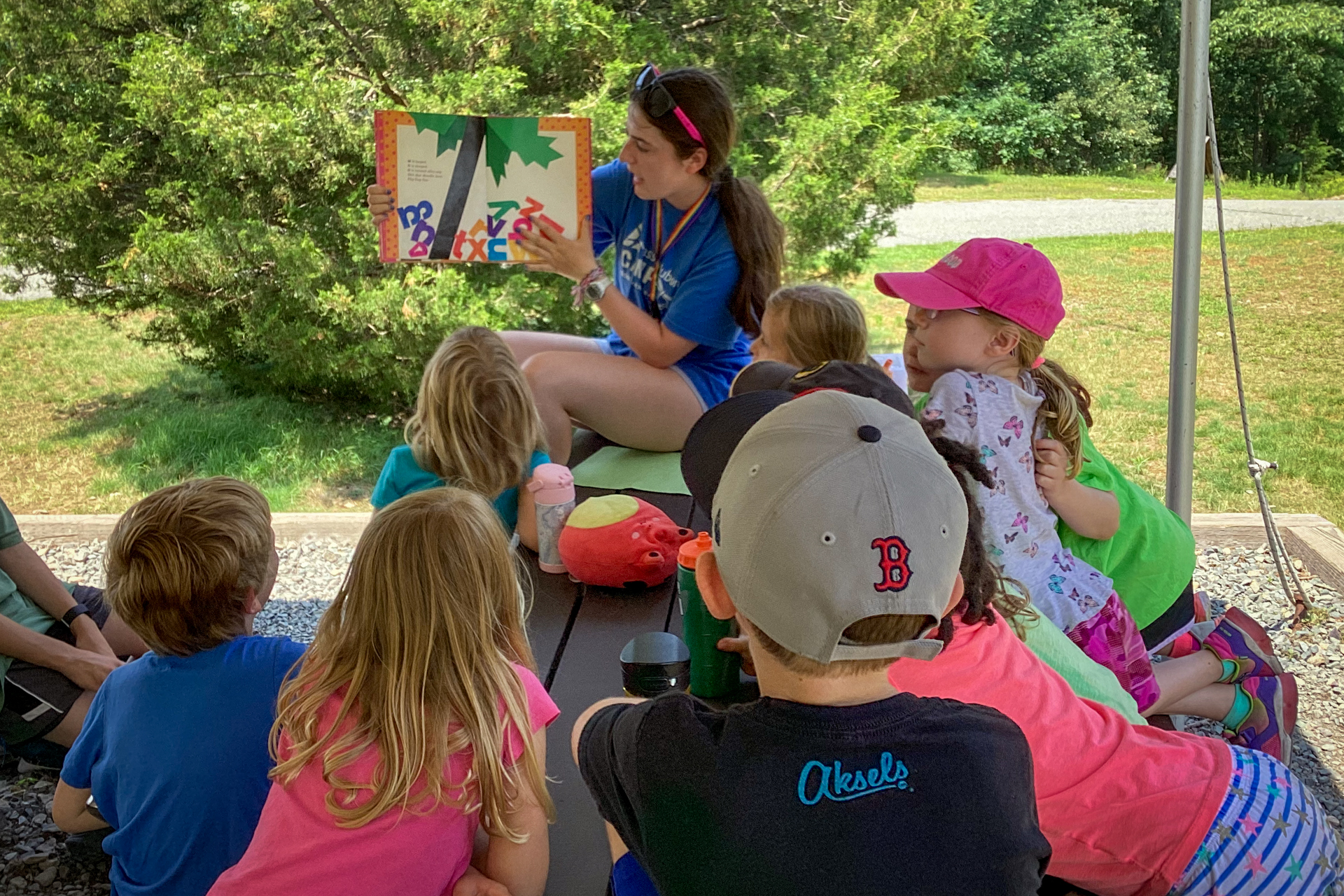 A Blue Hills Nature Camp counselor reads a picture book to a group of campers seated at an outdoor picnic table