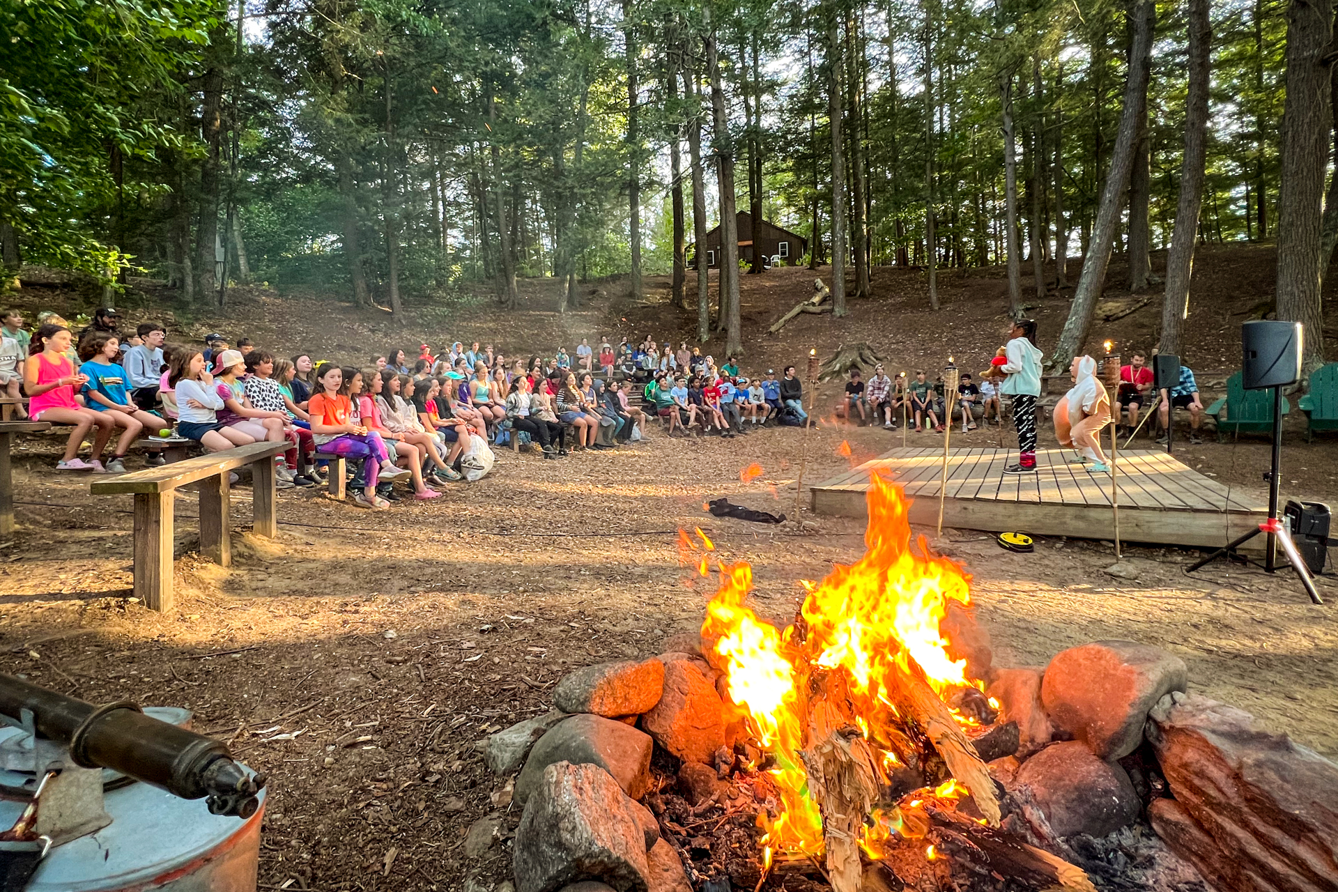 Two campers stand on the wooden stage in front of the outdoor amphitheater at Wildwood Camp, performing a skit to a large group of campers and staff. In the foreground, a campfire burns brightly.