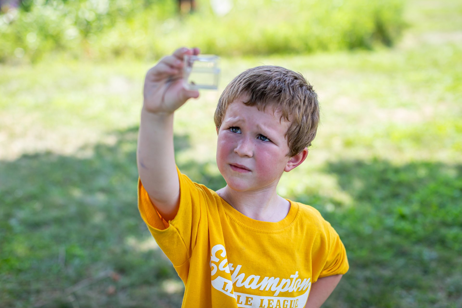 A camper wearing an Easthampton Little League shirt holds up a clear plastic specimen jar and peers at the insect inside