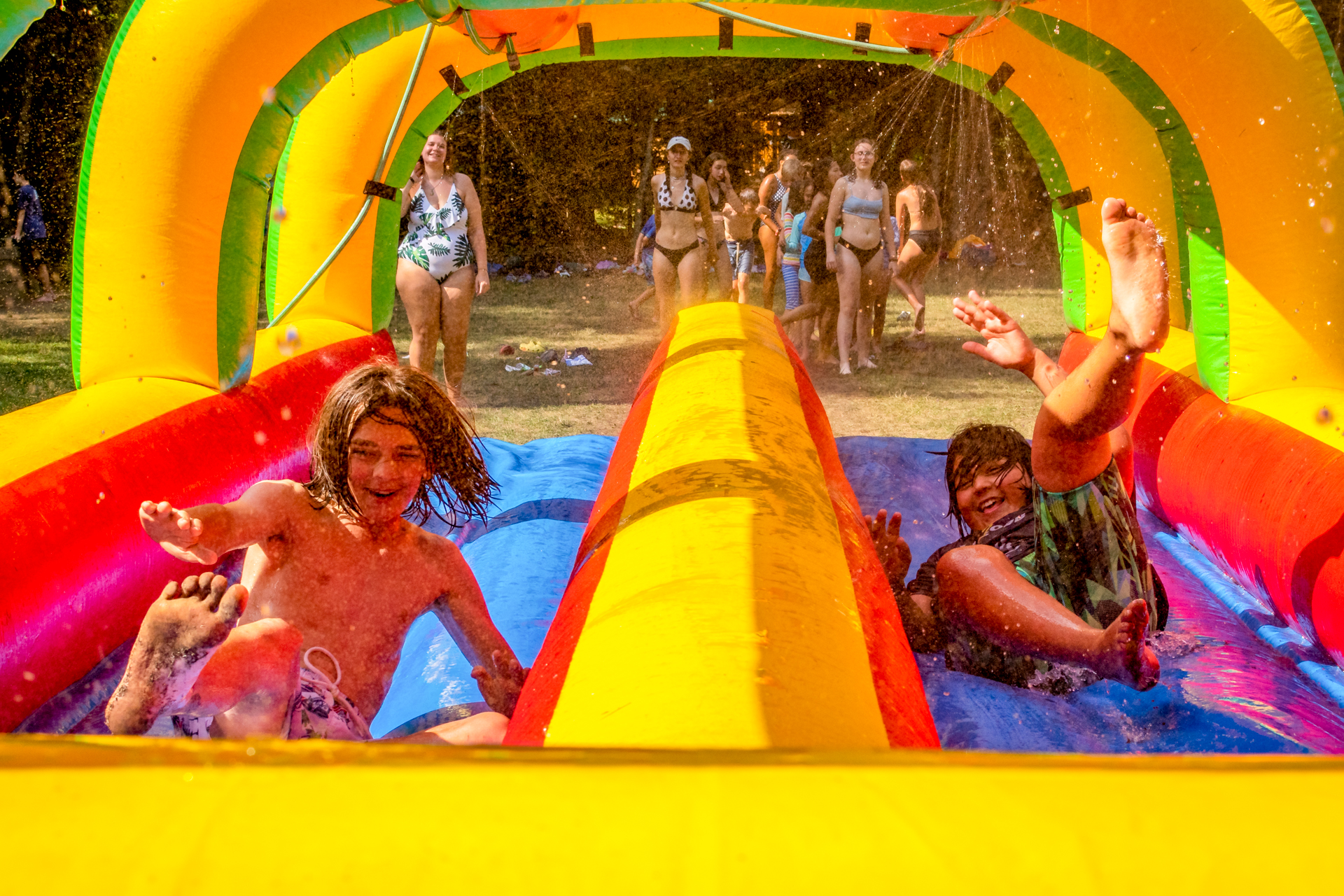 Two laughing campers are sliding down an inflatable waterslide with hoses spraying water down from above. In the background, a group of campers waits for their turn.
