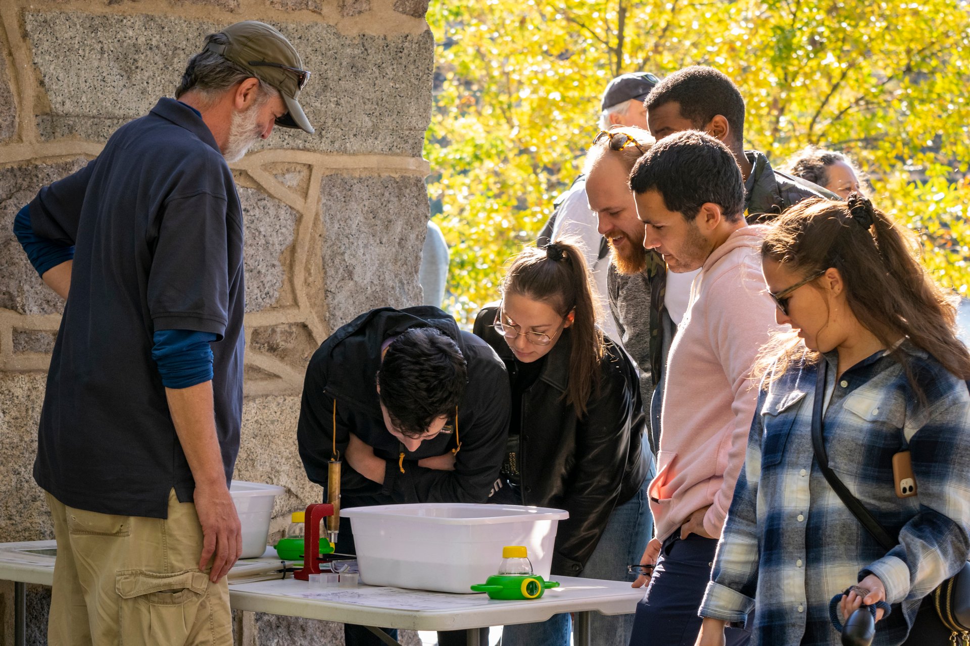 A man in a blue shirt and grey hat stands behind a table with two white buckets, and a small red microscope. A group of six people crowd around the table to examine the contents in the bucket.