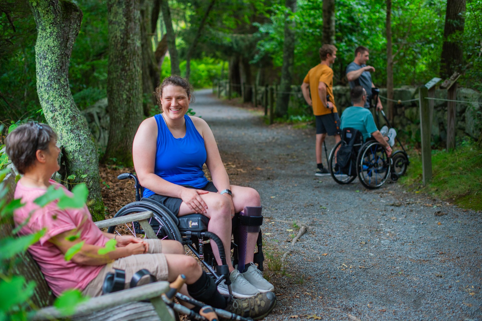 Two women at a bench beside a flat gravel path, one has bilateral crutches and the other is seated in a wheelchair, while a group of people in the background look at an interpretive sign along the accessible All Persons Trail at Stony Brook Wildlife Sanctuary