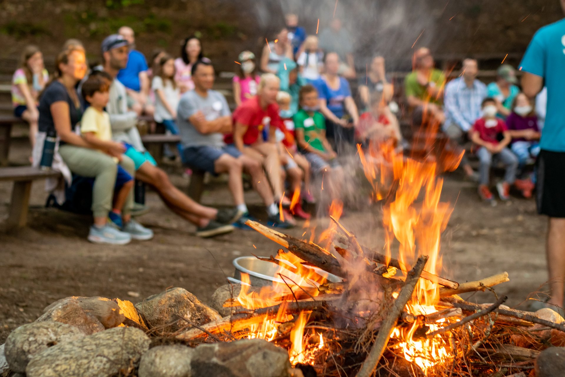 A campfire blazes in the foreground while in the background, slightly out of focus, a group of families sits together in the outdoor amphitheater at Wildwood, enjoying an evening Family Camp program