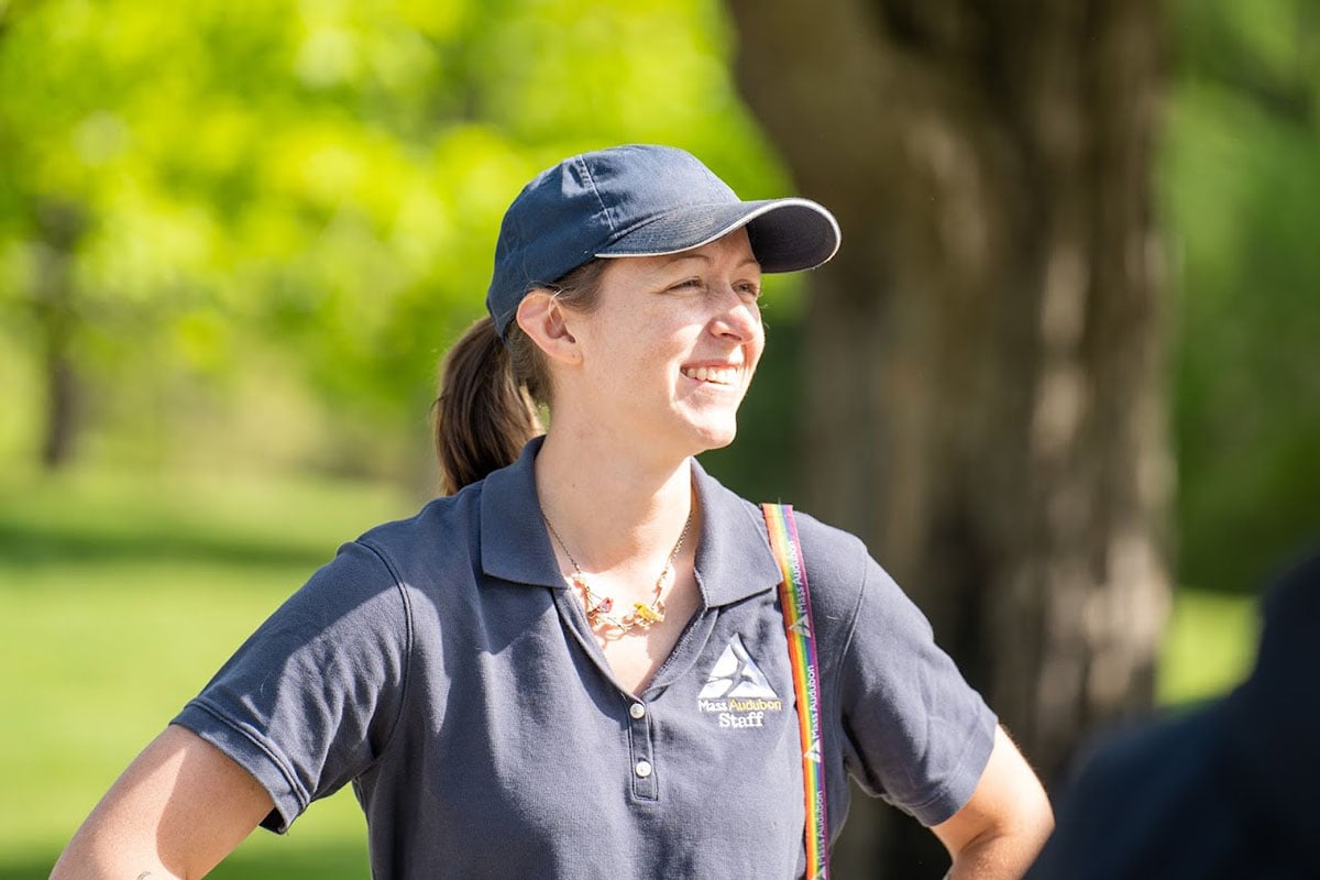 A woman in a Mass Audubon t-shirt and blue hat smiling in the distance.