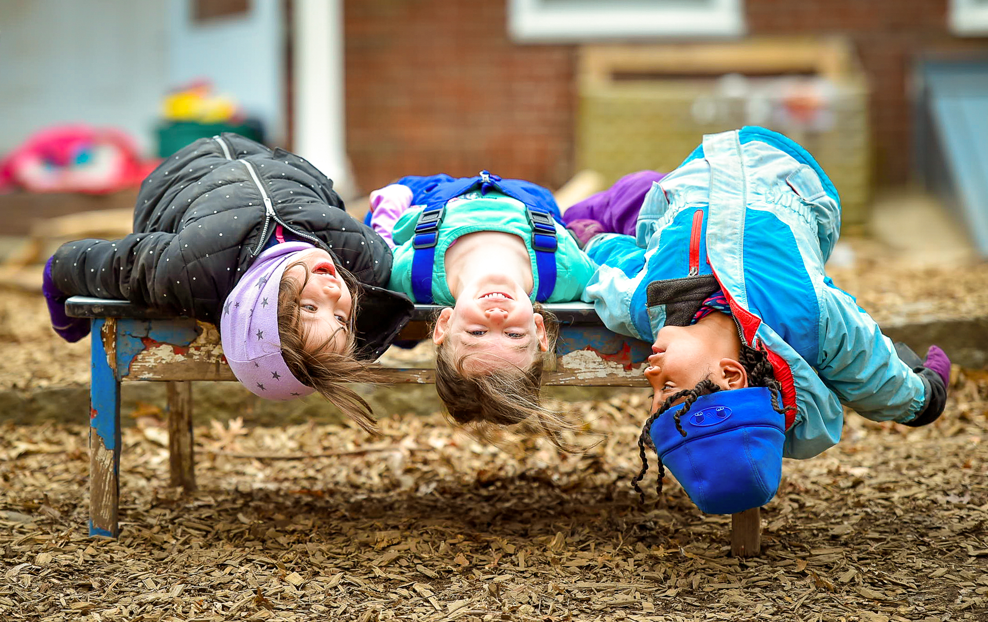 Three children laying on their backs, heads upside-down, on a playground bench at Drumlin Farm Community Preschool
