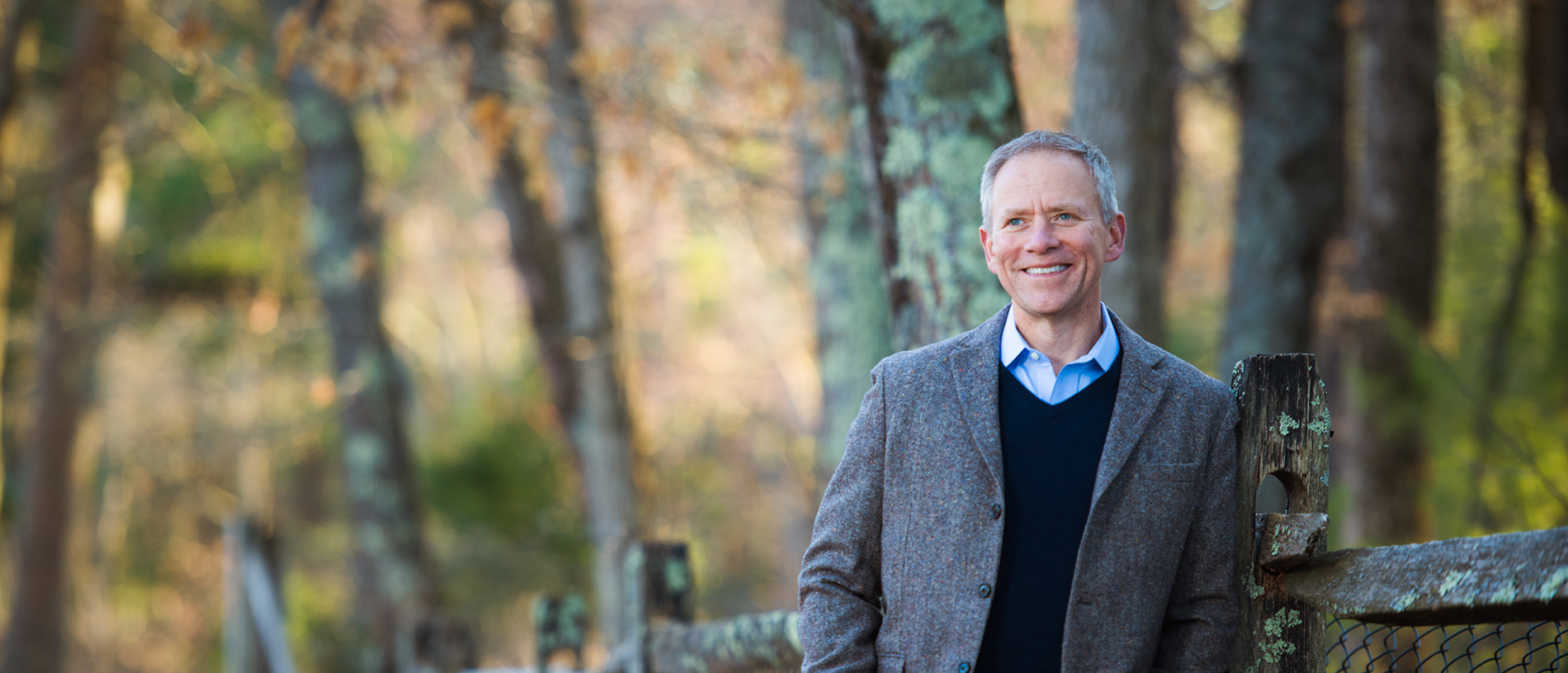 David O'Neill leaning against a wood fence wearing an open blazer, dark sweater, and blue collared shirt.