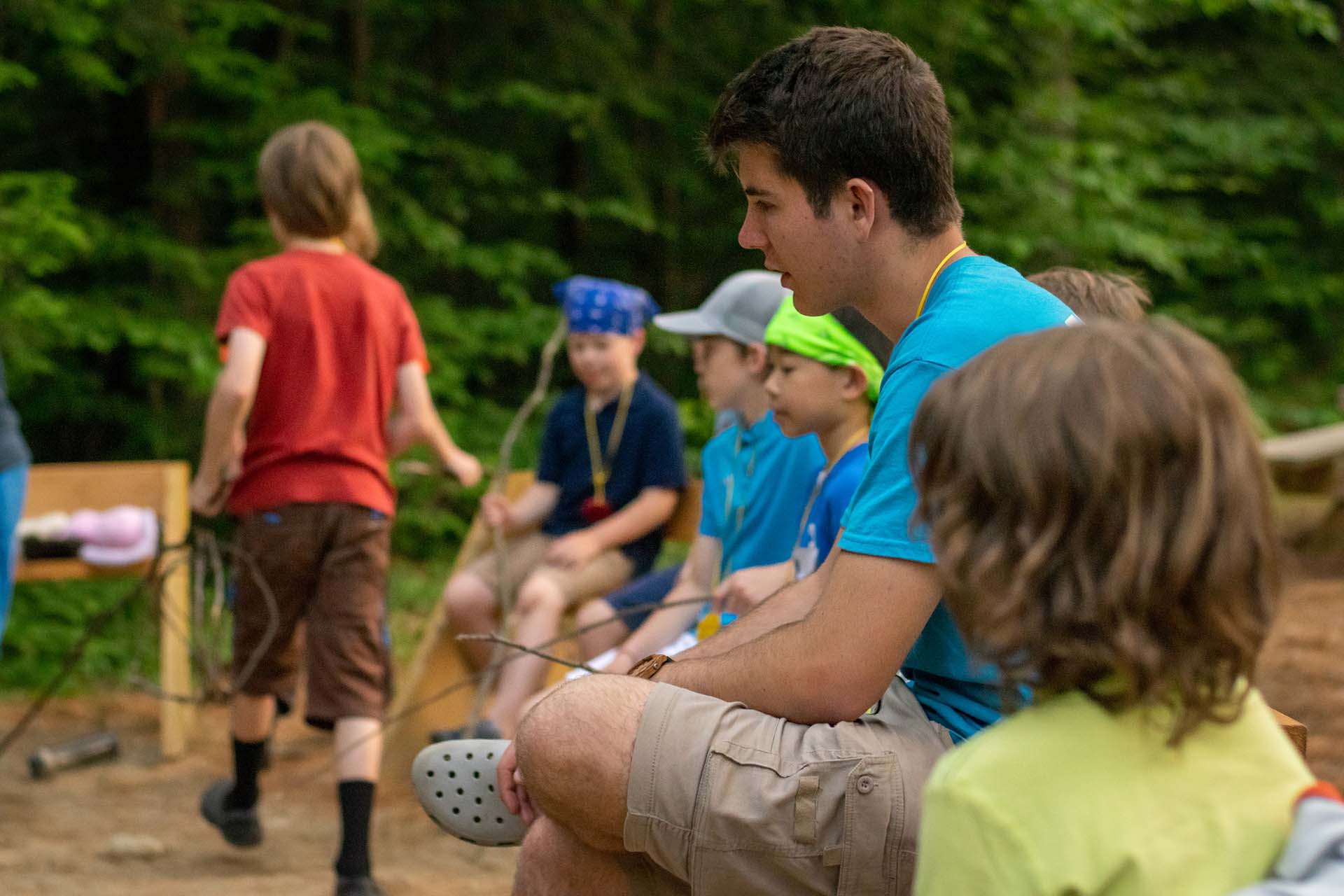 A man in a blue shirt sitting with kids, holding a stick.