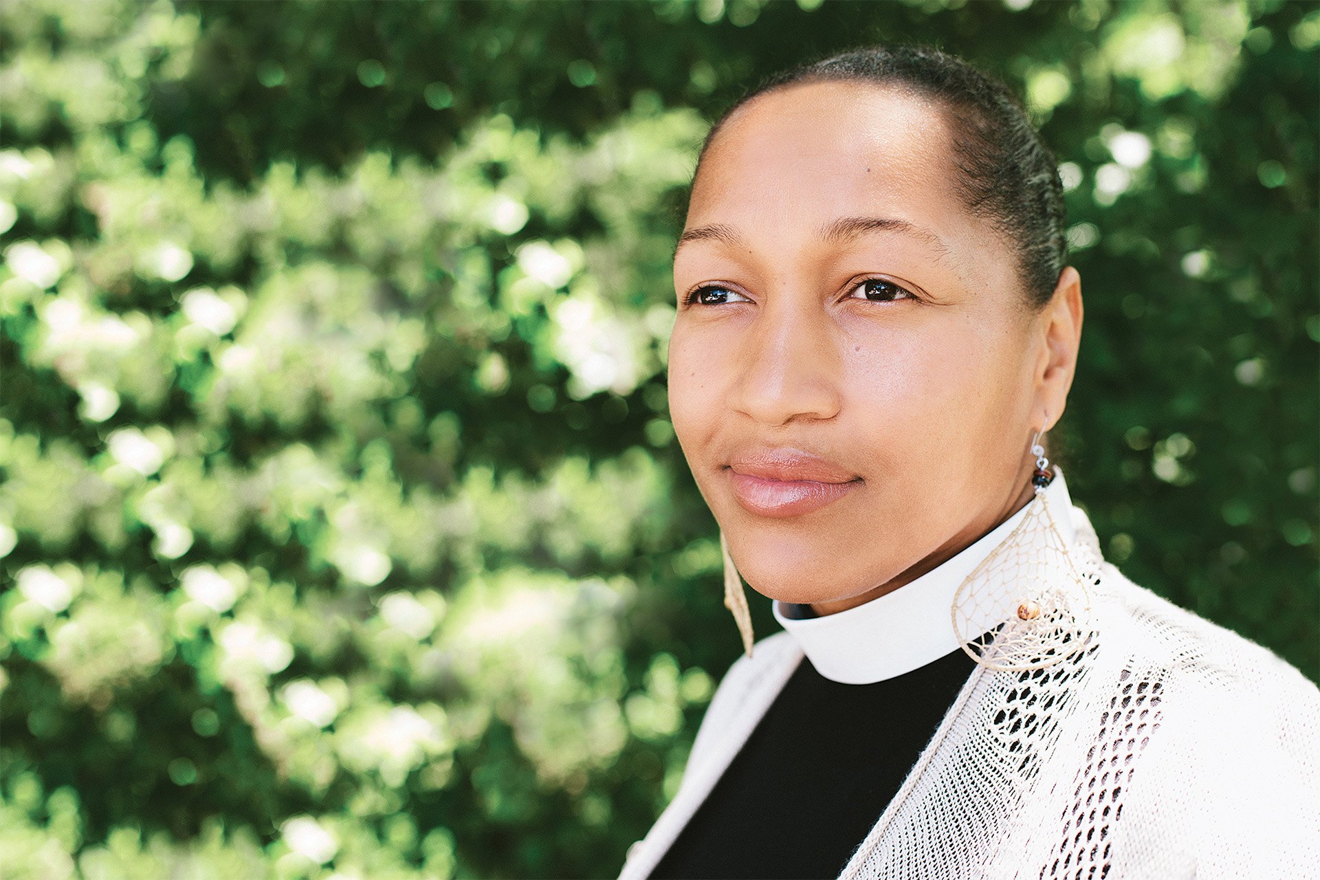A portrait of Rev. Mariama White-Hammond standing in front of a green, leafy background