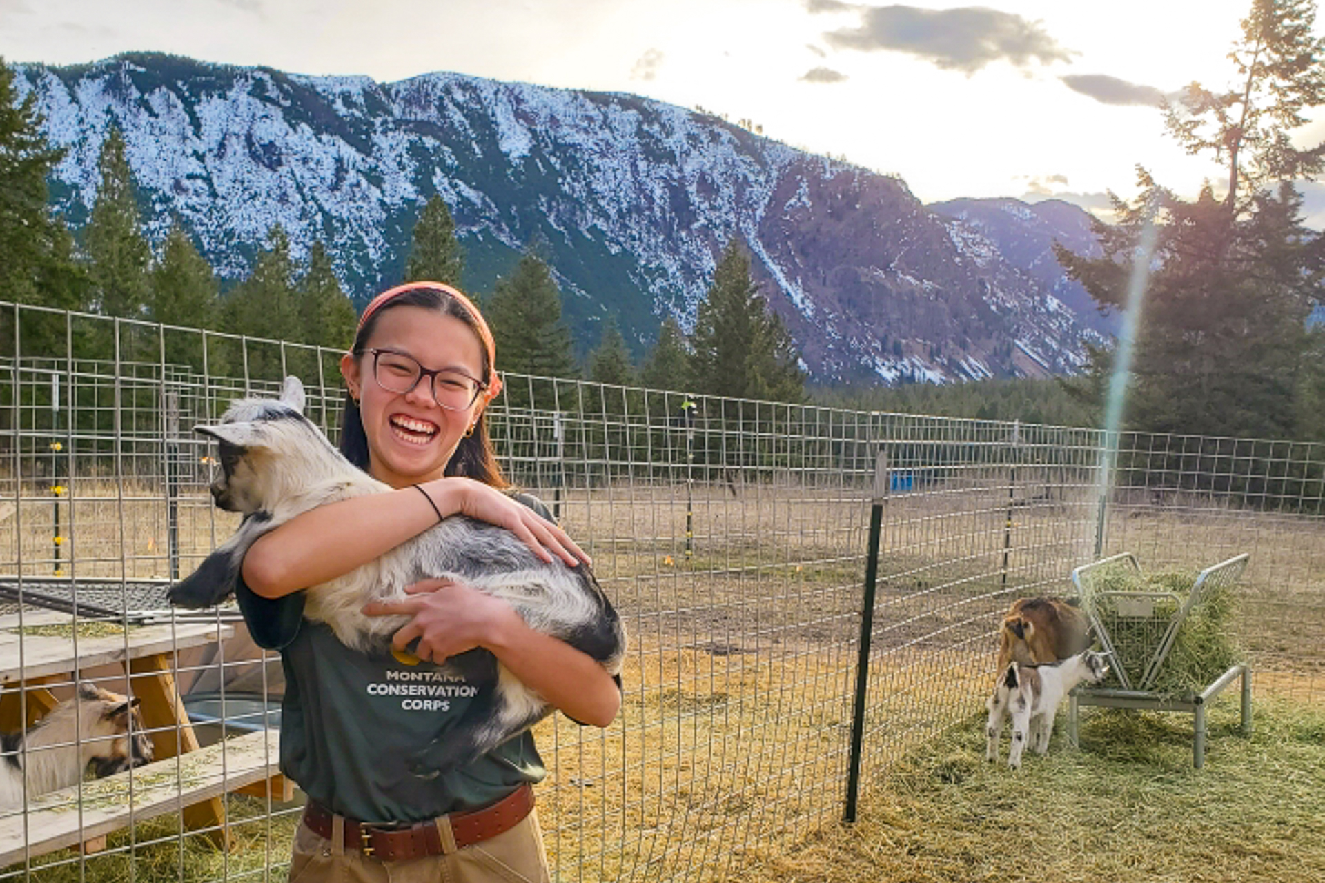 Mass Audubon's new coordinator for the Teen Adventure Trips program, Julia Drennan, stands inside a fenced-in enclosure with beautiful snow-dusted mountains in the background, She is holding a goat in her arms and smiling and is wearing a t-shirt for the Montana Conservation Corps.