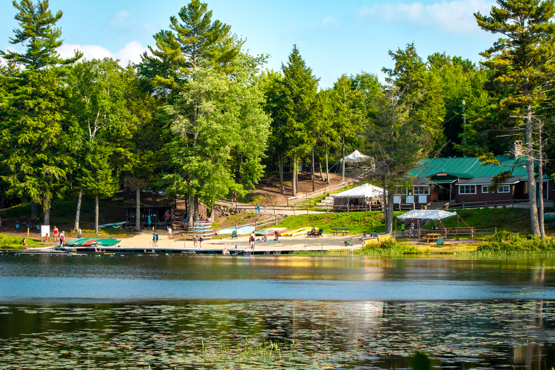 Looking back at the waterfront and dining hall at Wildwood Camp from across the water. A few campers can be seen on the beach, and the sun is shining through the green forest all around.