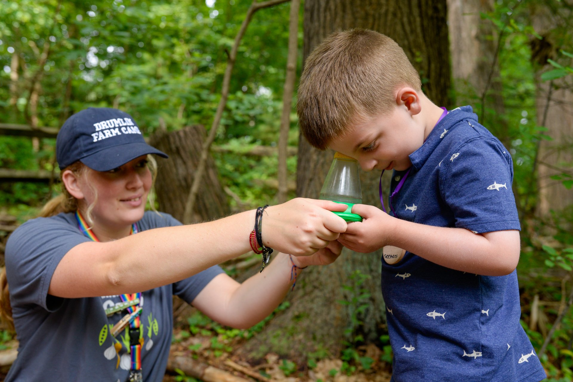 A counselor at Drumlin Farm Camp holds a magnifying bug catcher for a camper who is peering inside