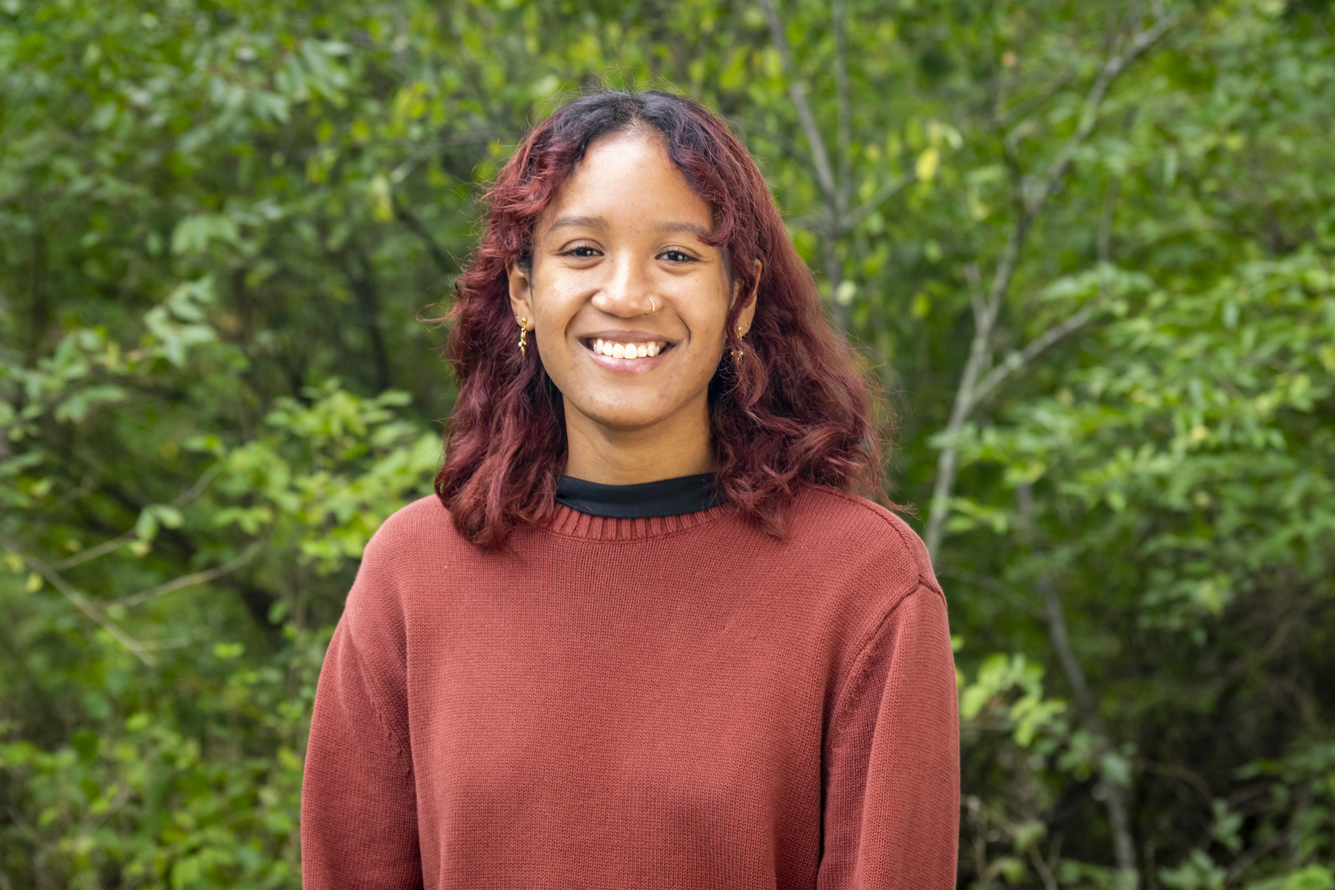 Isabella wearing a red sweater, smiling with green leaves in the background.