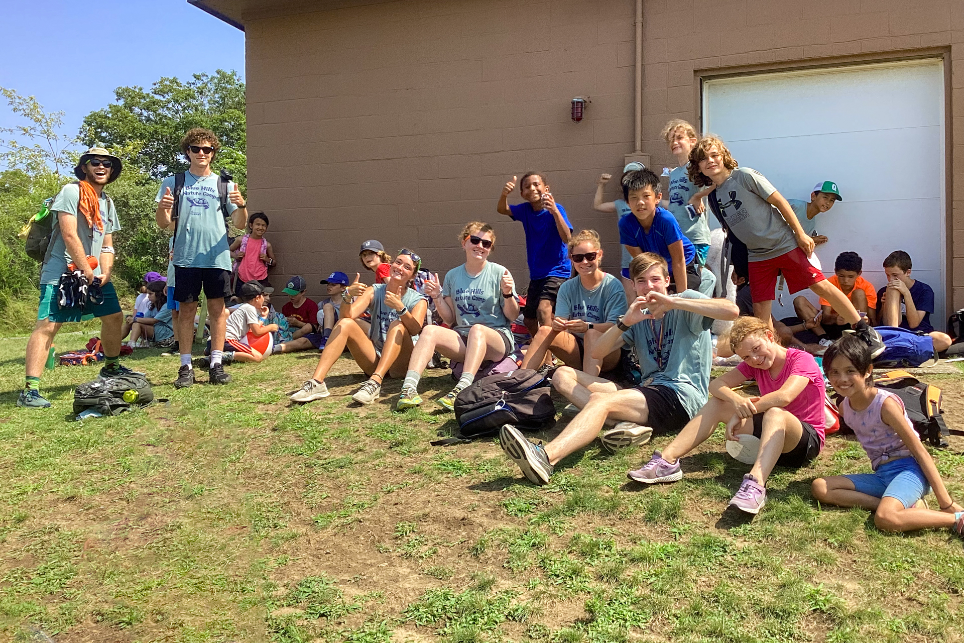 A group of smiling Blue Hills campers and counselors pose in front of a cinderblock building