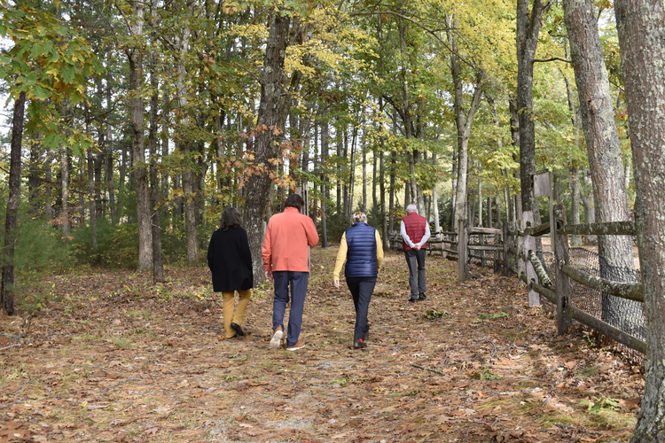 Four people walking on a bare trail. To their right is a wooden fence and a few trees, to their left is woods and shrubs.