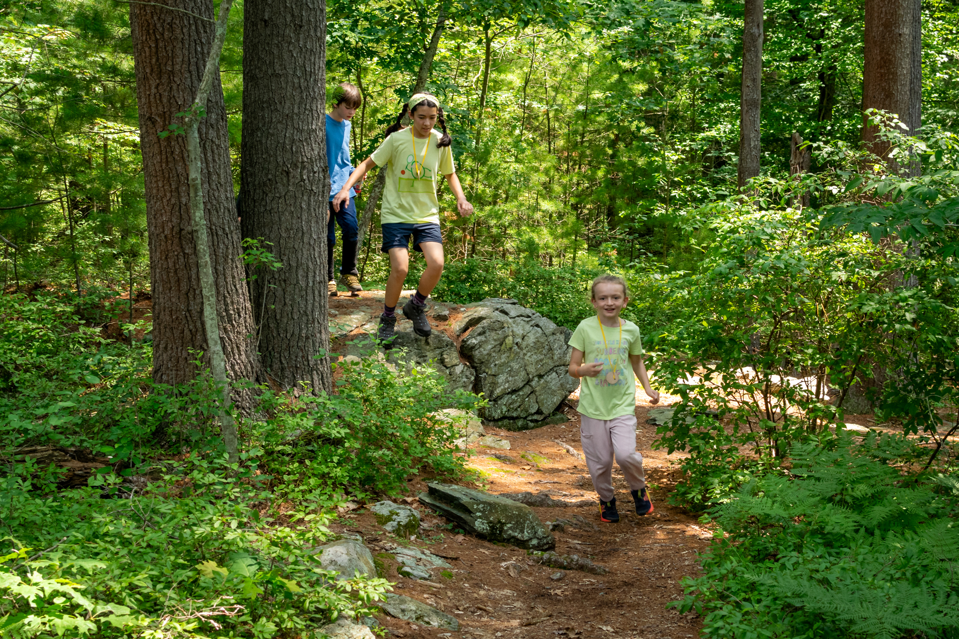 Three smiling Broadmoor campers running along a rocky trail through the forest
