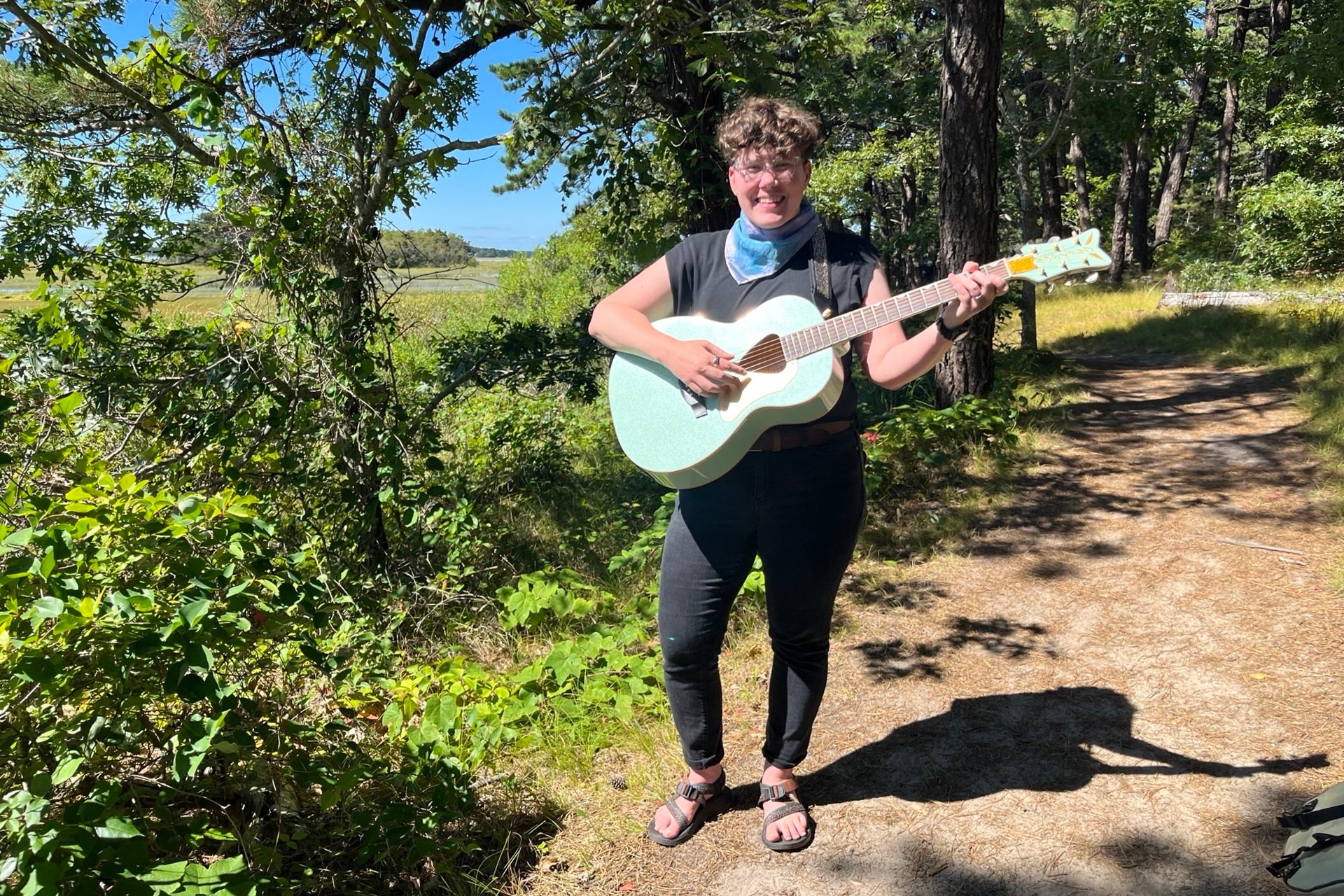 Person with short curly hair holding a light blue guitar on a trail
