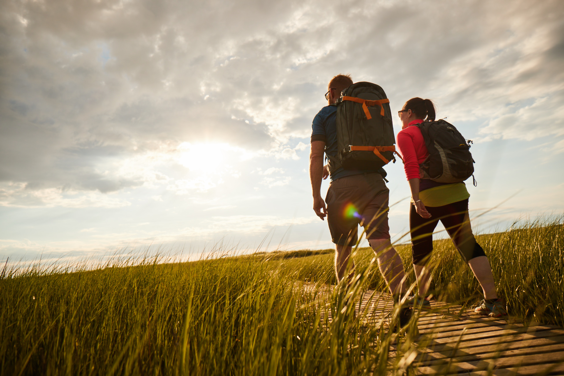 Two adults walking on a boardwalk at sunset, one pointing at something in the distance.