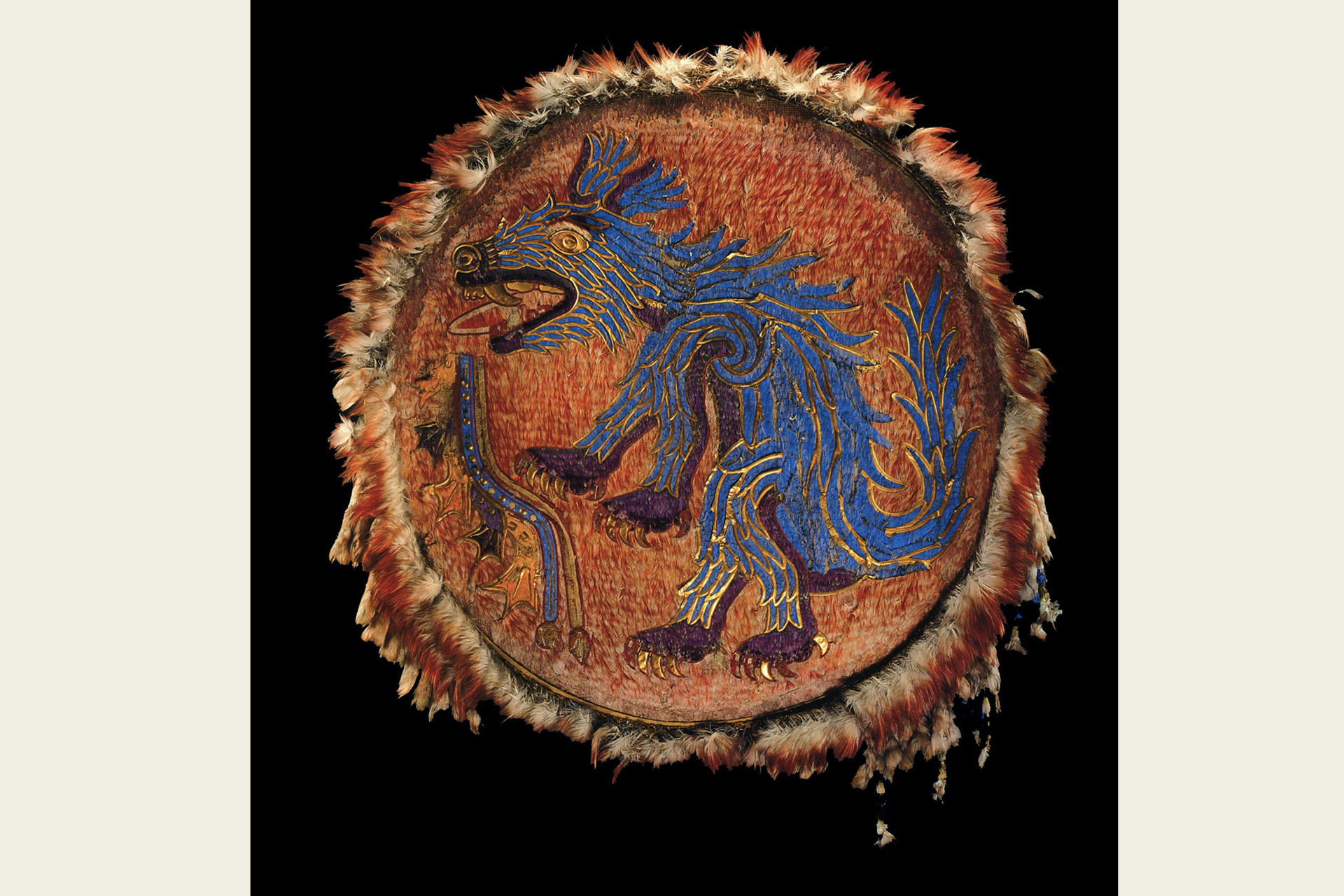 A museum display of a feather shield with a mosaic deisgn of a blue canid at its center