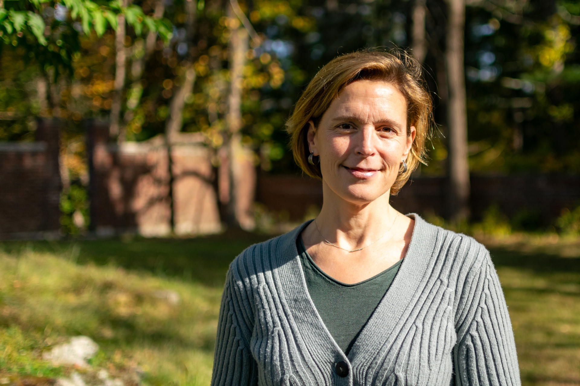 Woman with short hair smiling at the camera. Green grass and leaves are in the background.