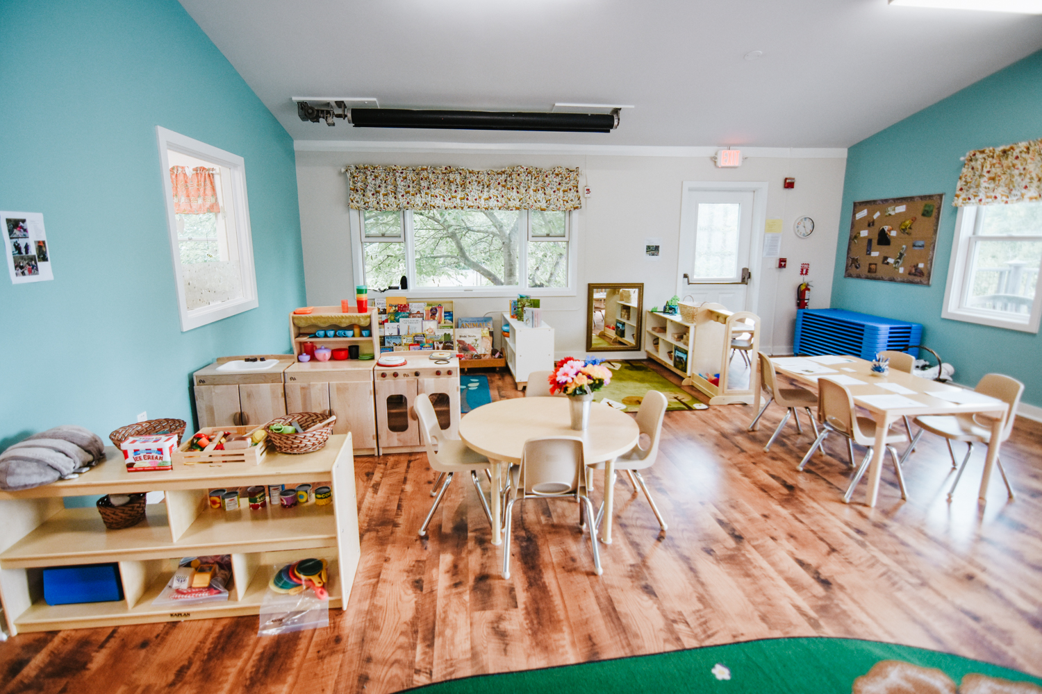 A view of the classroom at Stony Brook including two tables with chairs, play kitchen, blue walls, wood floor