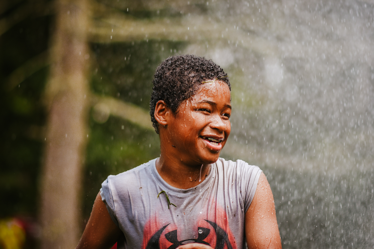 A smiling camper at Moose Hill Nature Camp wearing wet clothes and being showered by water from a firetruck hose on a hot day