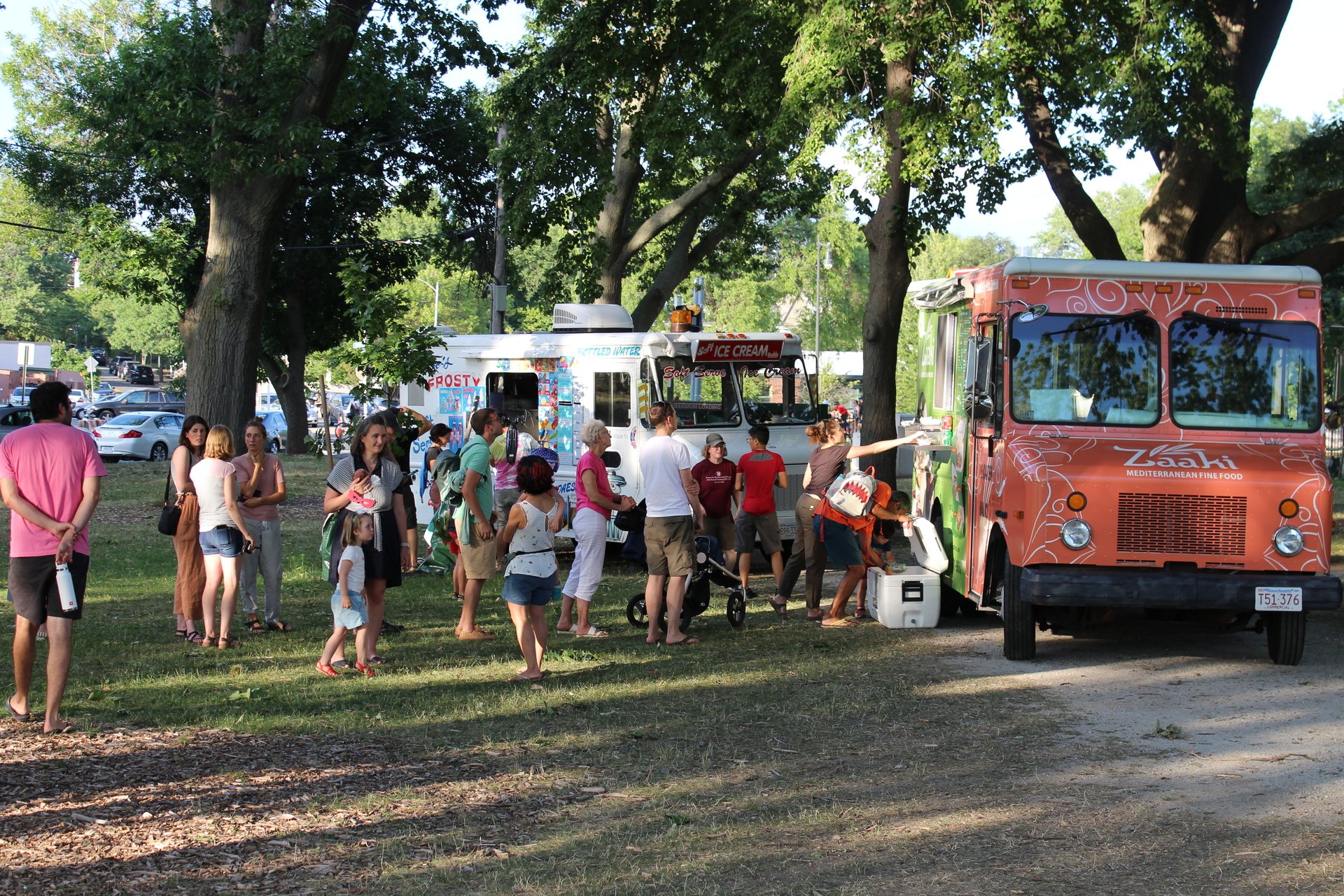 Two food trucks, one white and one orange, parked in the grass with lines of people waiting to order.