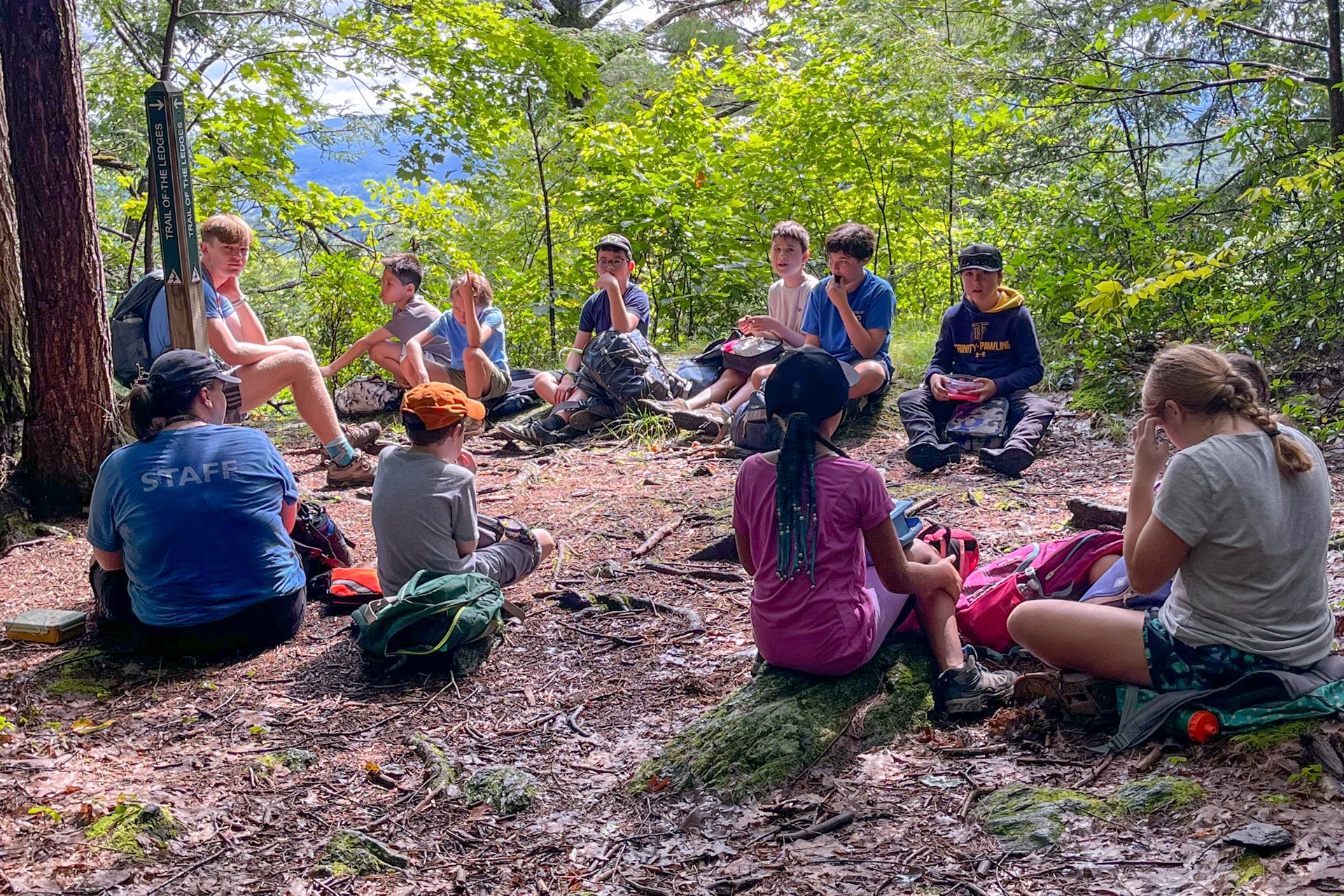 A group of Berkshire Nature Camp campers seated in a circle, enjoying a trail lunch on a hike to Trail of the Ledges at Pleasant Valley Wildlife Sanctuary