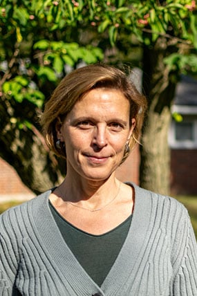 Woman with short hair smiling at the camera. Green grass and leaves are in the background.