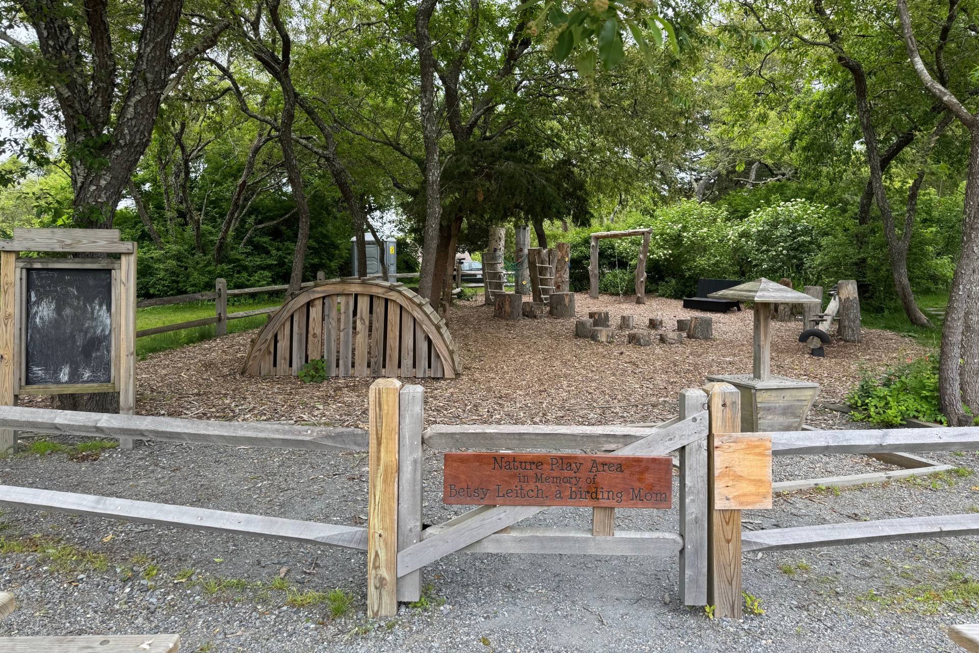 Long Pasture's Nature Play Area features wooden structures behind fence with a gate. Surrounding the area are green trees.
