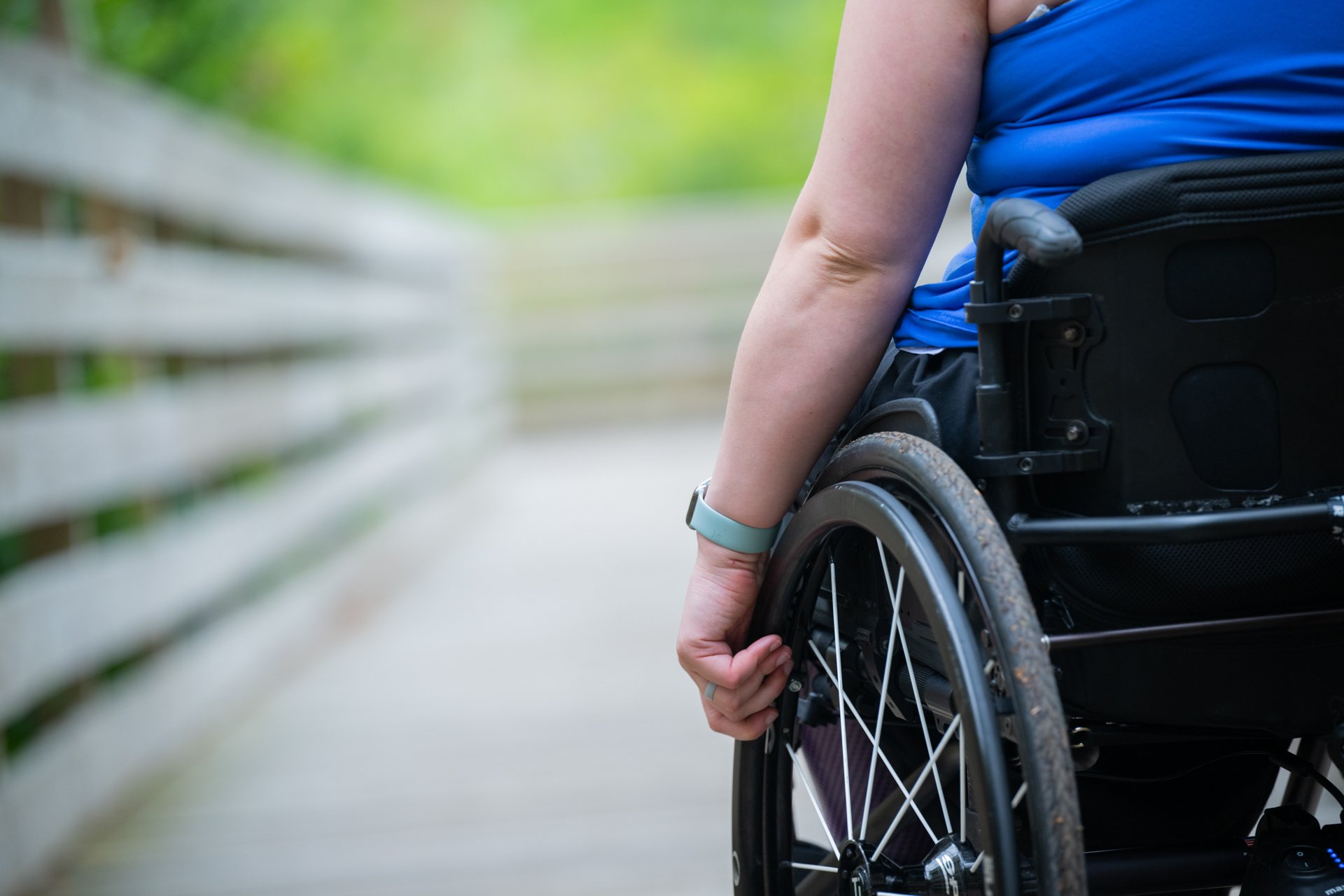 A close-up, cropped photo of a person seated in a wheelchair with their hand on the wheel, using the accessible All Persons Trail boardwalk