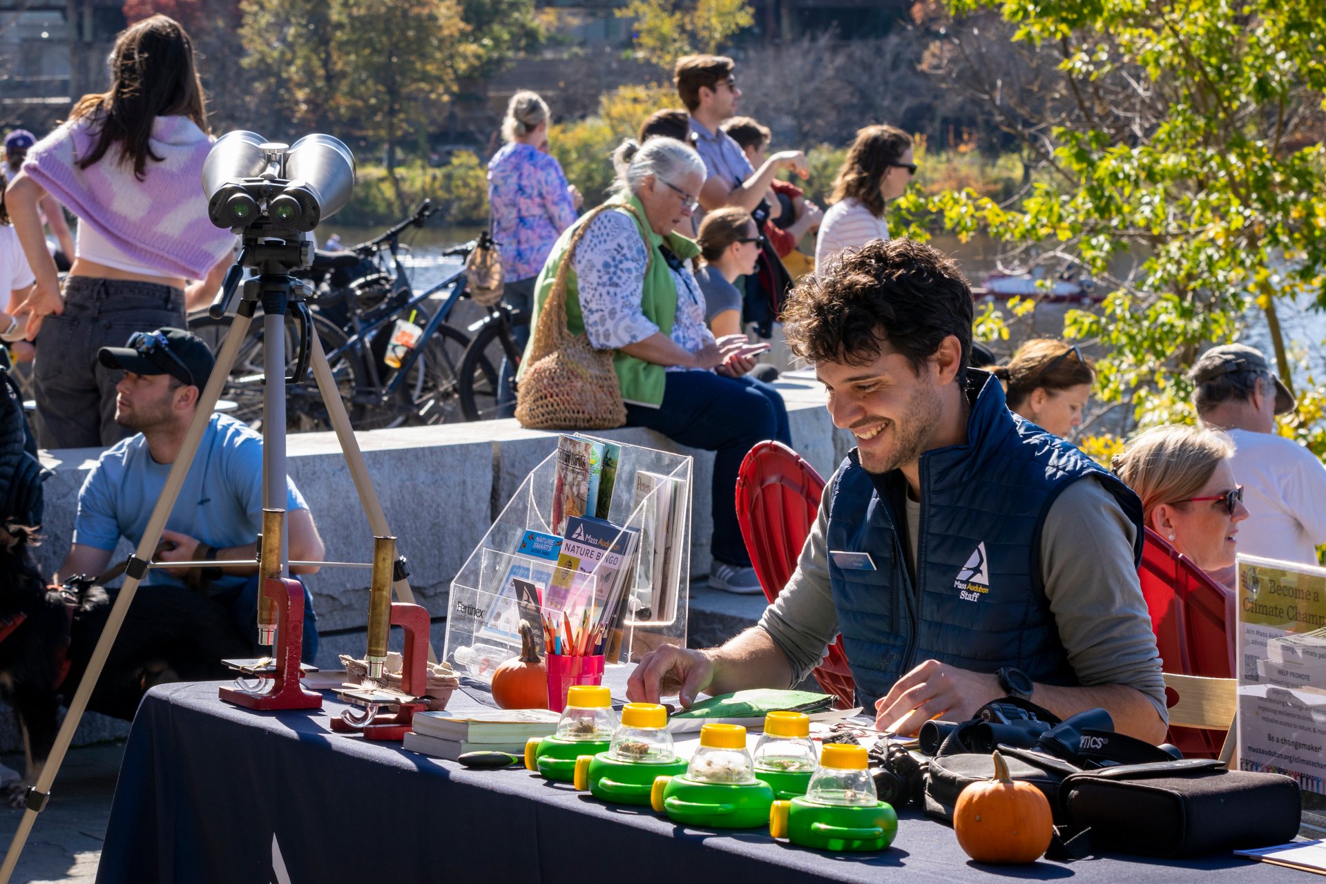 A man smiling with a Mass Audubon vest siting at a table with binoculars, microscopes, Mass Audubon promotional packets and paper.
