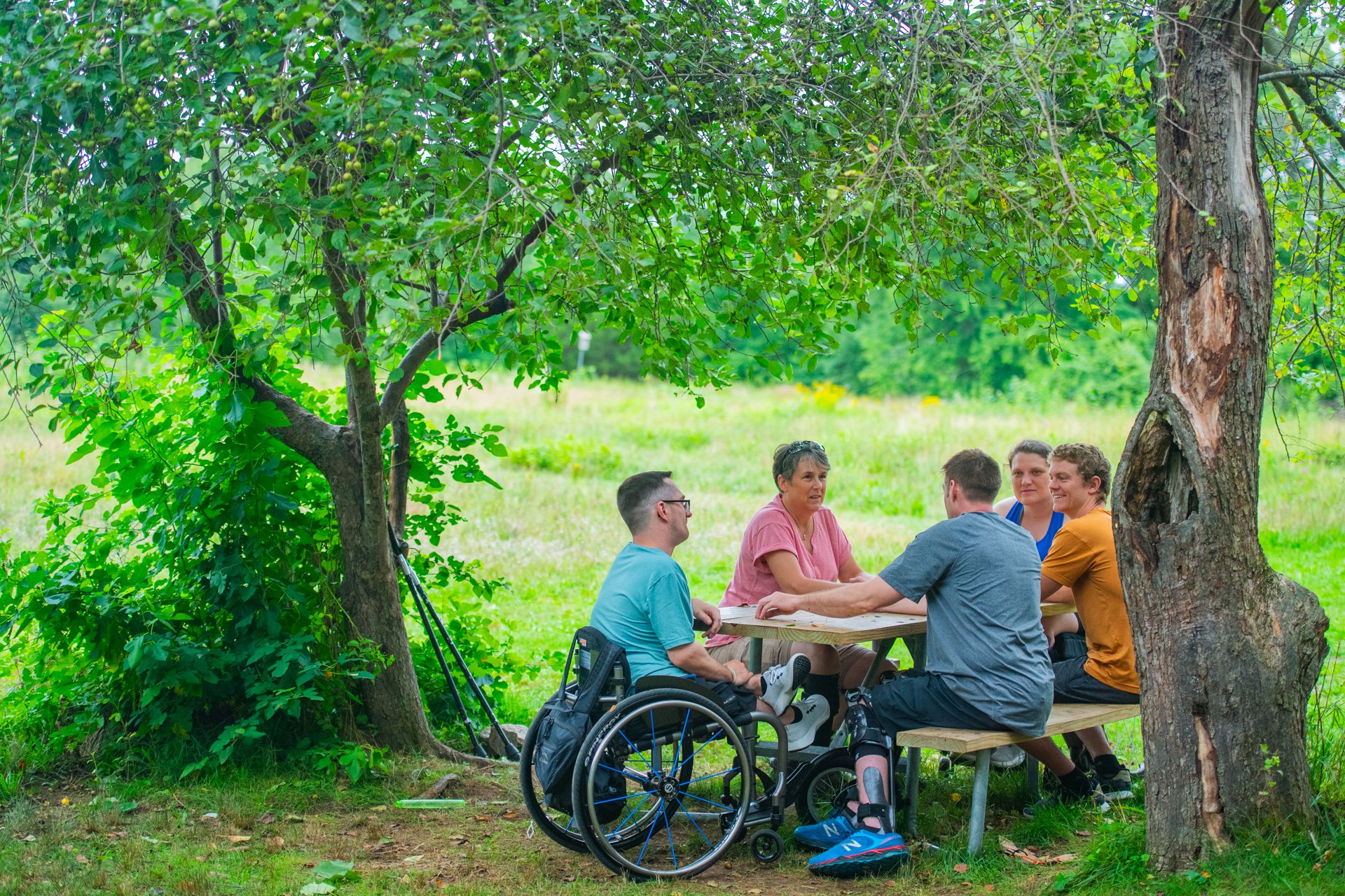 A group of five people, including one wheelchair user, are seated at an accessible picnic table beneath a tree, talking and smiling