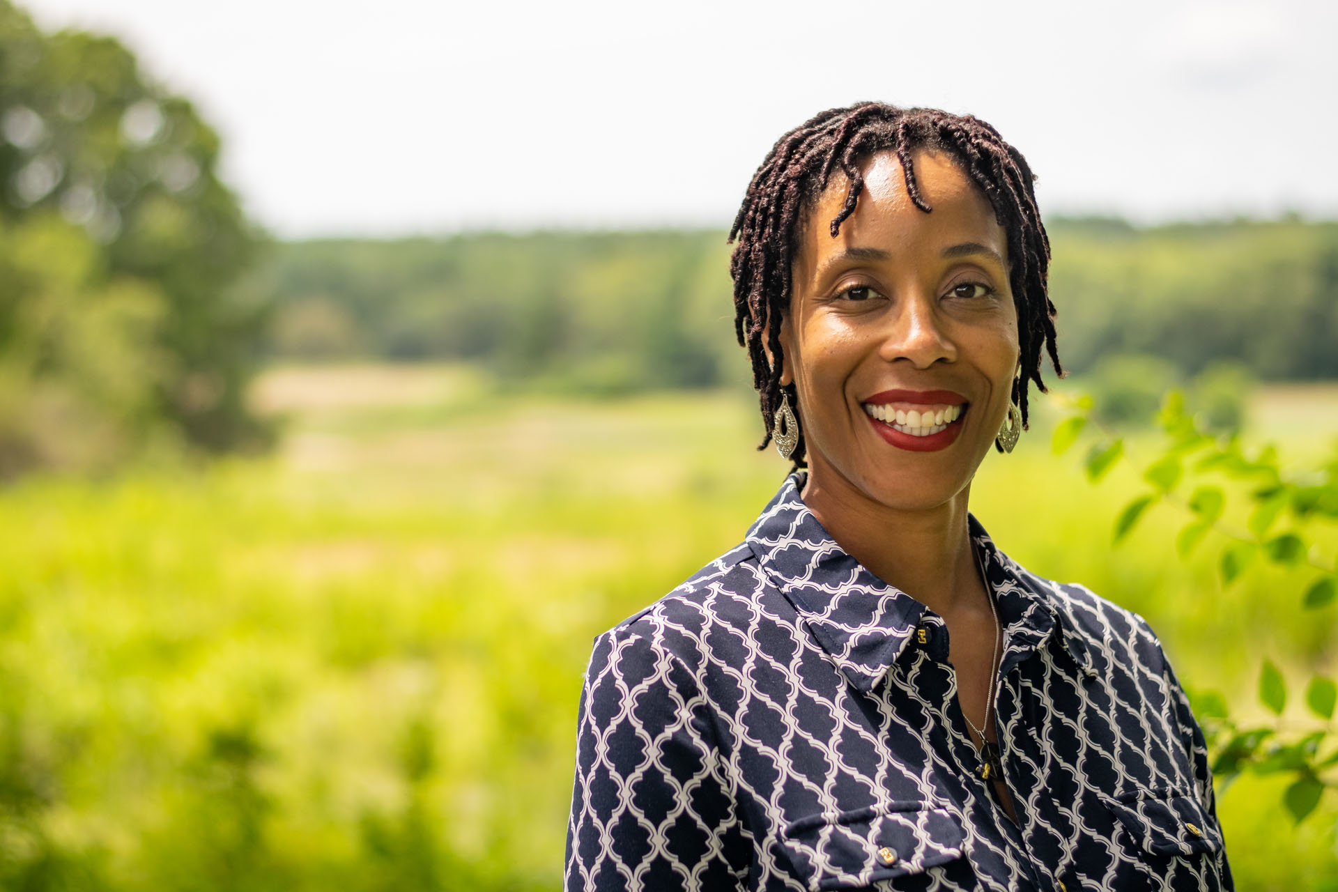 Woman smiling with a blue and white shirt with a green pasture in the background.